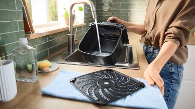 Persona che risciacqua un cestello nero per friggitrice ad aria sotto l'acqua corrente in un moderno lavello da cucina con paraschizzi piastrellato di verde.