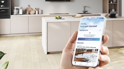 Smartphone portatile che mostra un'app per la domotica in una cucina moderna con mobili leggeri e pavimento in legno.