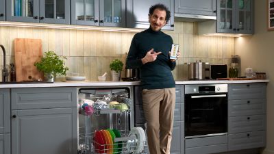 Uomo in una cucina moderna accanto a una lavastoviglie aperta, mentre mostra uno smartphone con un'app di controllo