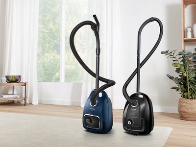 Two Bosch bagged cylinder vacuums stand on a cream rug in a light, bright living room.