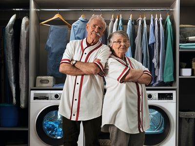 Two fit retirees in matching white tops stand back to back in an elegant laundry room.
