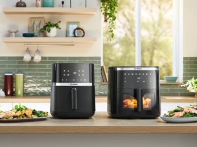 Dois modelos de Air Fryer da Bosch posicionados lado a lado num balcão de cozinha luminoso. Vários pratos e tigelas de comida, incluindo pão de ervas, frango, assado e batatas fritas, são colocados ao lado dos aparelhos, mostrando pratos confecionados com as Air Fryers.