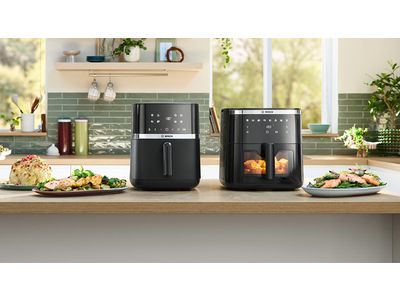 Two Bosch Air Fryer models positioned side by side on a bright kitchen counter. Several plates and bowls of food, including herb bread, chicken, roast, and fries, are placed next to the appliances, showcasing dishes made using the Air Fryers.