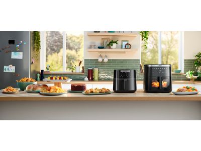 Two Bosch Air Fryer models positioned side by side on a bright kitchen counter. Several plates and bowls of food, including herb bread, chicken, roast, and fries, are placed next to the appliances, showcasing dishes made using the Air Fryers.