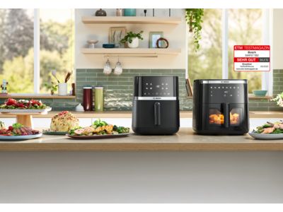 Zwei Modelle der Bosch Heißluftfritteuse nebeneinander auf einer hellen Küchenarbeitsplatte. Mehrere Teller und Schüsseln mit Lebensmitteln – darunter Kräuterbrot, Hähnchen, Braten und Pommes frites – stehen neben den Geräten und enthalten Gerichte, die mit der Heißluftfritteuse zubereitet wurden. Das größere Gerät ist mit dem neuen ETM Award ausgezeichnet.