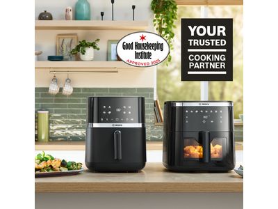 Two Bosch Air Fryer models positioned side by side on a bright kitchen counter. Several plates and bowls of food, including herb bread, chicken, roast, and fries, are placed next to the appliances, showcasing dishes made using the Air Fryers.