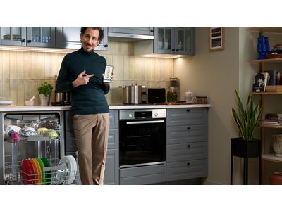 Uomo in cucina con lavastoviglie aperta, stoviglie colorate all'interno, mentre tiene un dispositivo in mano.