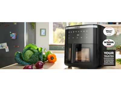 Two Bosch Air Fryer models positioned side by side on a bright kitchen counter. Several plates and bowls of food, including herb bread, chicken, roast, and fries, are placed next to the appliances, showcasing dishes made using the Air Fryers.