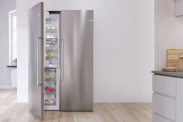 Réfrigérateur argent side-by-side pose-libre Bosch dans une cuisine blanche. Porte ouverte avec vue sur des provisions.