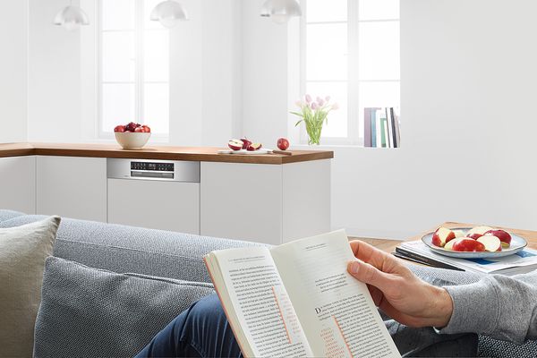  Person quietly reads on sofa while a Bosch dishwasher runs nearby. 