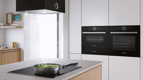 Una cocina para el cocinero ambicioso: una placa de inducción integrada en una isla de cocina moderna con salteado de brócoli en una sartén y hornos gemelos a la altura del pecho en el fondo