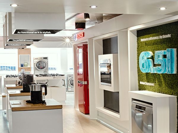 Erősen megvilágított Bosch bemutatóterem, amelyben vízforraló, mosogatógép, beépített sütő és piros hűtőszekrény található