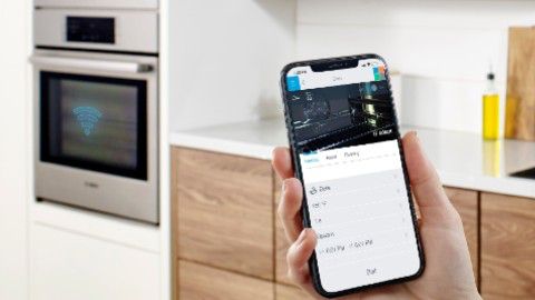 iPhone negru care controlează un cuptor cu abur încorporat într-o bucătărie albă cu dulapuri din lemn în partea de jos