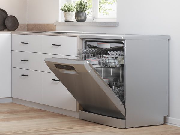 Rozsdamentes acél szabadonálló mosogatógép, kissé nyitott ajtóval, egy ablakpárkány alatt, amelyen friss zöldfűszerek vannak