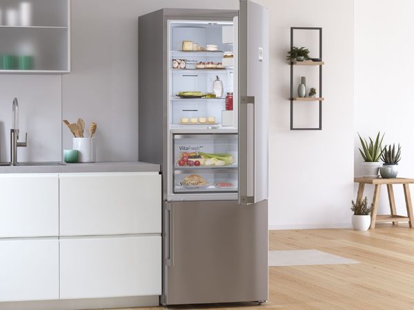 	 Open stainless steel fridge filled with delicious pastries and fresh produce in a white and grey kitchen with frosted upper cabinets and aloe and desert plants in the background 
