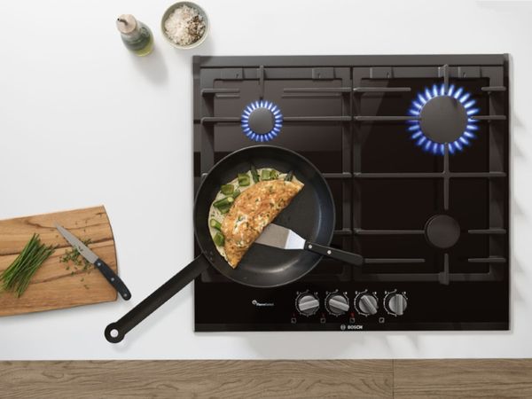 Black ceramic 4-burner gas hob with lit flames and a spatula ready to flip a green bell pepper and asparagus omelette. Next to the frying pan are freshly cut chives on a cutting board