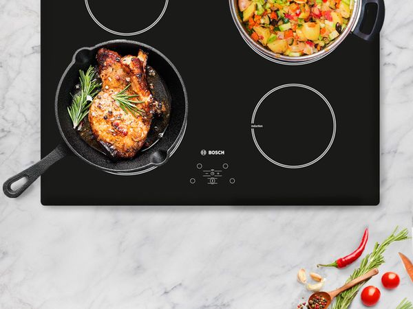 Colourful veggies and rosemary chicken cooking on a black induction hob mounted on a marble kitchen counter