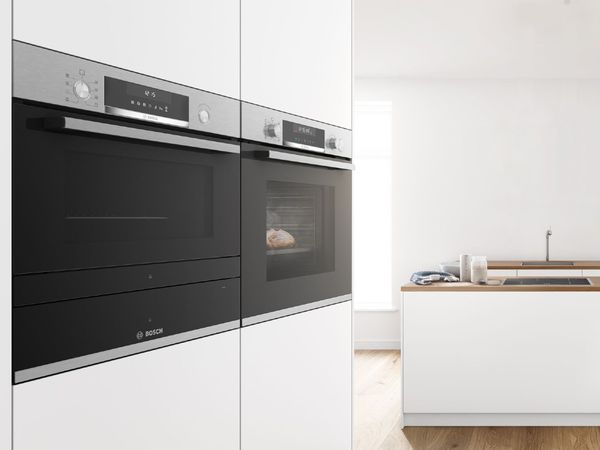 Two black built-in wall ovens with stainless steel bar handles in an all-white kitchen with an island, sink and hob in the background