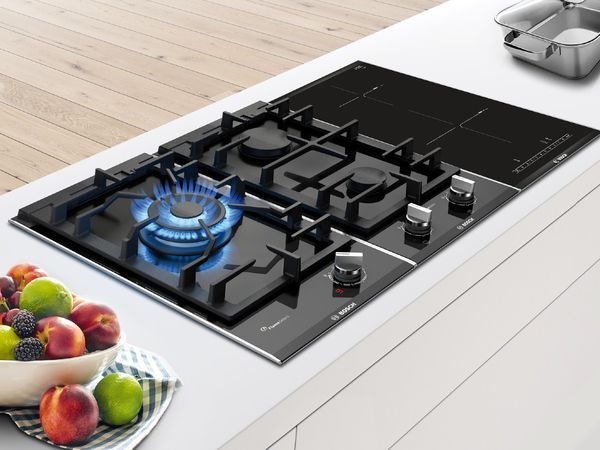 Trio of black domino hobs with a lighted gas burner on a white kitchen counter in between a fresh fruit bowl and steel cookware