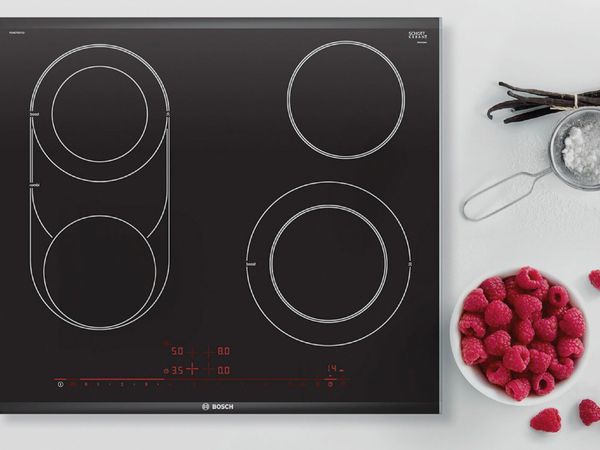 White kitchen counter with a black electric hob next to a bowl of red raspberries, a bundle of fresh vanilla and a small sieve of powdered sugar