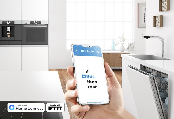 Smartphone avec l'application IFTTT (If this then that) devant une cuisine blanche moderne avec lave-vaisselle, machine à café et four Bosch. 