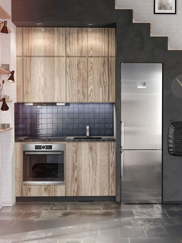Маленькая L-образная кухня под лестницей с дубовыми шкафами, холодильником и духовкой из нержавеющей стали. Холодильник и стиральная машина встроены в стену лестницы.