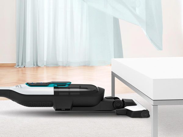 A cordless vacuum cleans under a coffee table. The curtains waft in the background.