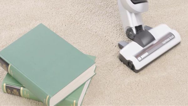 Прахосмукачка почиства килим. До нея има книги.