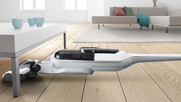 A cordless vacuum cleans under a living room table. There is a couch in the background.