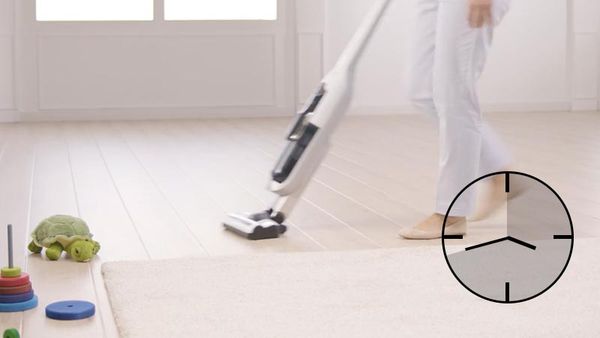 A woman vacuums, there are toys in the corner. A clock symbol is placed on the photo.