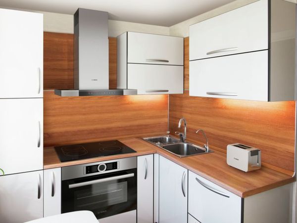 Небольшая, уютная Г-образная кухня с классическими белыми шкафами и столешницей из дерева теплых оттенков, переходящей в облицованную деревом заднюю панель