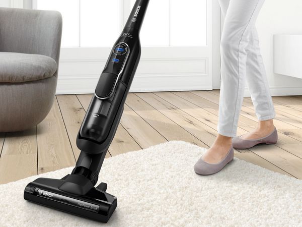 A black cordless vacuum cleaner vacuums a beige carpet. A grey sofa in the bachground.