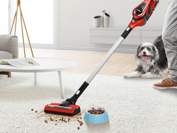 A cordless vacuum cleaner vacuums dog food from the carpet. A dog is in the background.