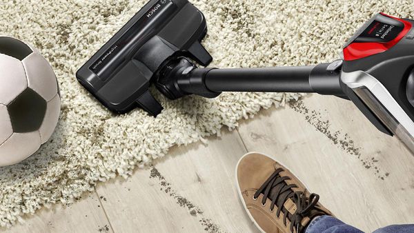 A cordless vacuum cleans the carpet, you see a foot and a football next to it.