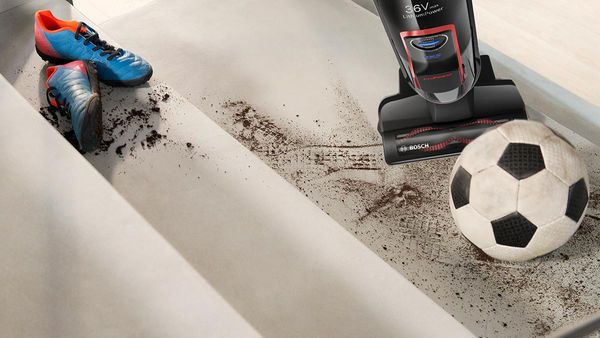 A cordless vacuum cleans the carpet, you see a foot and a football next to it.