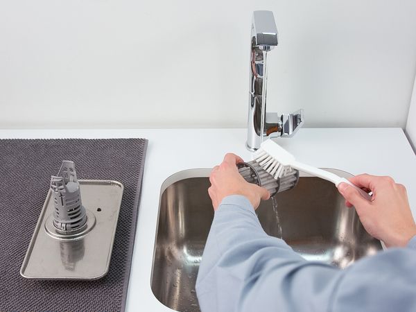 Deux mains tenant une brosse et une unité de filtrage d'un lave-vaisselle Bosch sous l'eau courante