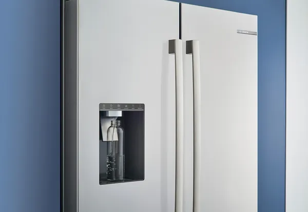 Un réfrigérateur contemporain avec un distributeur d'eau devant un mur bleu, montrant une bouteille en verre près de la zone de distribution.