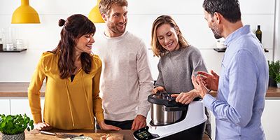 Vier Personen stehen vor einer Kücheninsel mit Holzoberfläche. In der Mitte von ihnen auf der Kücheninsel steht ein Cookit, dessen Deckel eine Frau mit grauem Pullover öffnet.
