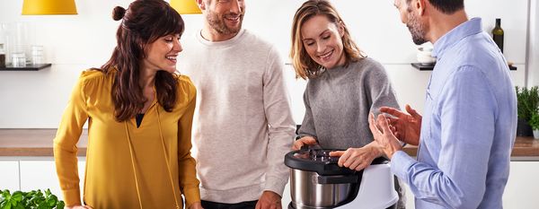 Vier Personen stehen vor einer Kücheninsel mit Holzoberfläche. In der Mitte von ihnen auf der Kücheninsel steht ein Cookit, dessen Deckel eine Frau mit grauem Pullover öffnet.