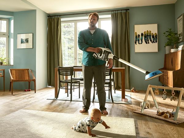 גבר עומד בסלון עם שואב אבק Unlimited של Bosch בידו. תינוק זוחל על שטיח לפניו.
