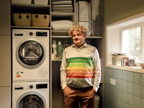 Мъж, в перално помещение със сушилня с термопомпа Bosch. Усмихва се, защото енергийно ефективната сушилня с термопомпа пести енергия благодарение на най-ефективния енергиен етикет, който също е нарисуван на пуловера му.