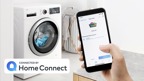 Bosch washer in a modern bathroom, hand in foreground with smartphone open to Home Connect app