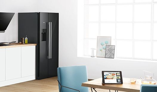Modern black fridge in a bright kitchen, next to a table with a tablet and decorative items. 
