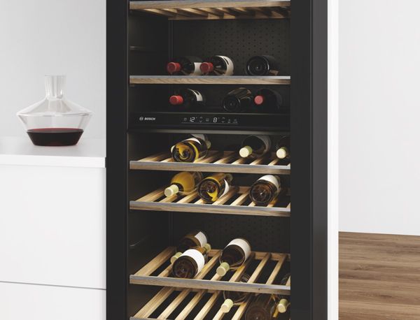 Carafe remplie de vin rouge corsé posée sur un comptoir à côté d'une cave à vin pose libre équipée de six étagères, d'une porte en verre, d'un éclairage LED et d'une collection de vins rouges et blancs.