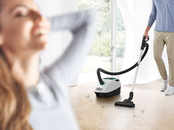 Smiling woman taking a deep breath as a man vacuums with a Bosch ProSilence model in the background  