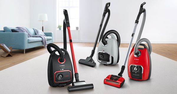 Three bagged vacuum cleaners in black, red and silver, forming a triangle on an off-white rug in a living room with hardwood flooring