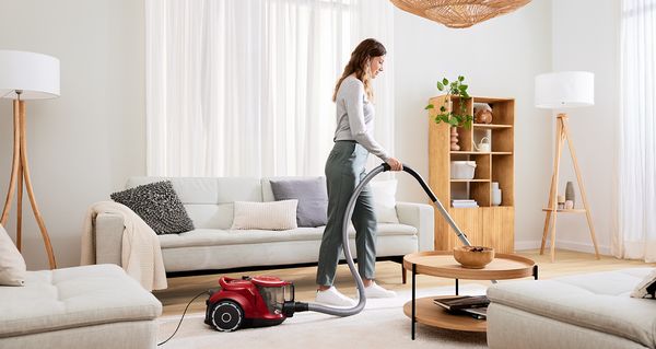A woman uses a canister vacuum to cleans a cream rug in a bright living room.