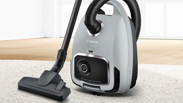 Compact grey cylinder vacuum on a white rug with floor-length windows in the background