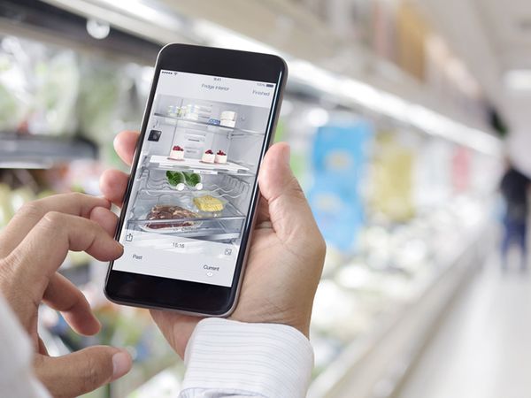 Mâini ținând un smartphone într-un supermarket. Pe ecran este interiorul frigiderului inteligent.