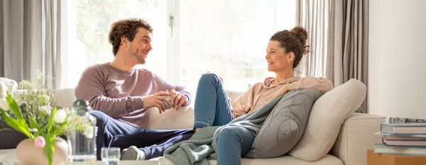 Un bărbat și o femeie stau pe o canapea și zâmbesc unul către celălalt.